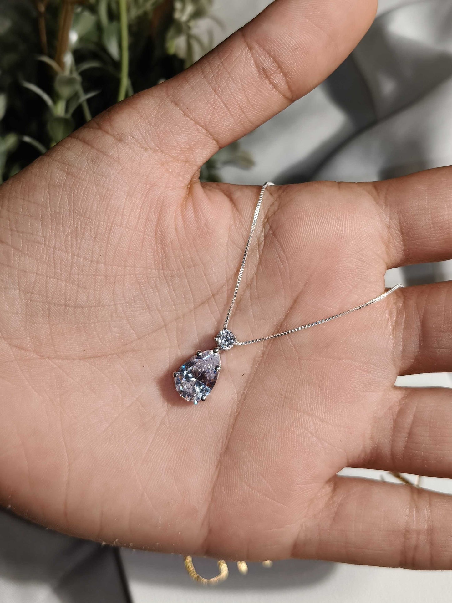 Teardrop-shaped pendant on a chain held in a hand against a neutral background